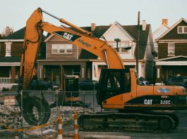 Excavator working in a suburban neighborhood construction site, with residential houses in the background.