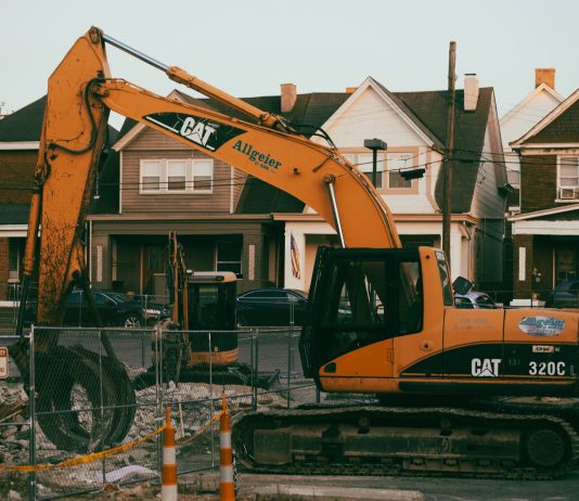 Excavator working in a suburban neighborhood construction site, with residential houses in the background.