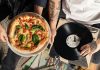 Overhead shot of a pizza and vinyl record held by tattooed individuals enjoying a casual indoor gathering.