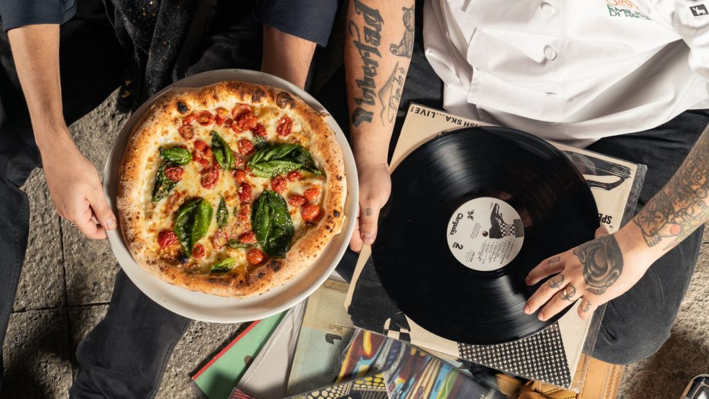 Overhead shot of a pizza and vinyl record held by tattooed individuals enjoying a casual indoor gathering.