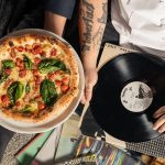 Overhead shot of a pizza and vinyl record held by tattooed individuals enjoying a casual indoor gathering.