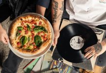 Overhead shot of a pizza and vinyl record held by tattooed individuals enjoying a casual indoor gathering.