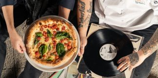 Overhead shot of a pizza and vinyl record held by tattooed individuals enjoying a casual indoor gathering.