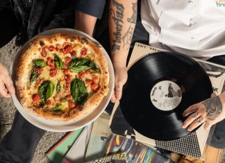 Overhead shot of a pizza and vinyl record held by tattooed individuals enjoying a casual indoor gathering.