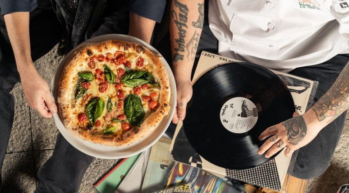 Overhead shot of a pizza and vinyl record held by tattooed individuals enjoying a casual indoor gathering.