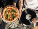 Overhead shot of a pizza and vinyl record held by tattooed individuals enjoying a casual indoor gathering.