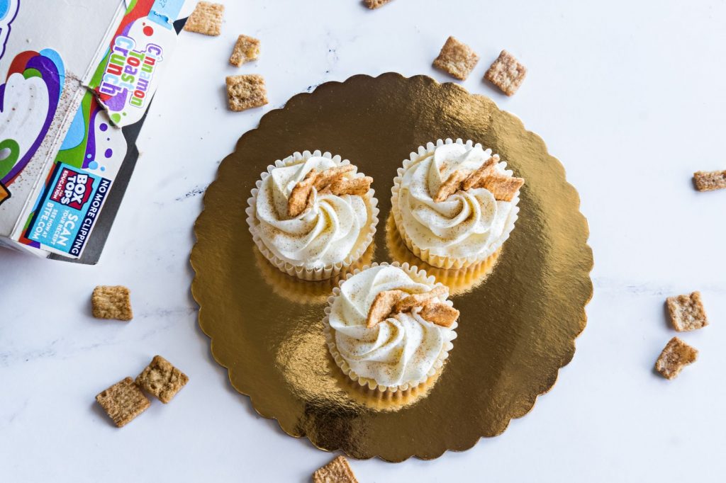 Top view of three cinnamon toast cupcakes with frosting on a golden tray, surrounded by cereal.