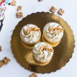 Top view of three cinnamon toast cupcakes with frosting on a golden tray, surrounded by cereal.