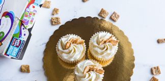 Top view of three cinnamon toast cupcakes with frosting on a golden tray, surrounded by cereal.