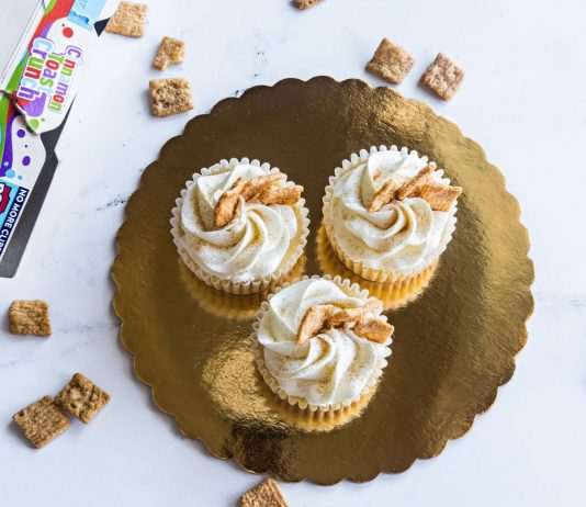 Top view of three cinnamon toast cupcakes with frosting on a golden tray, surrounded by cereal.
