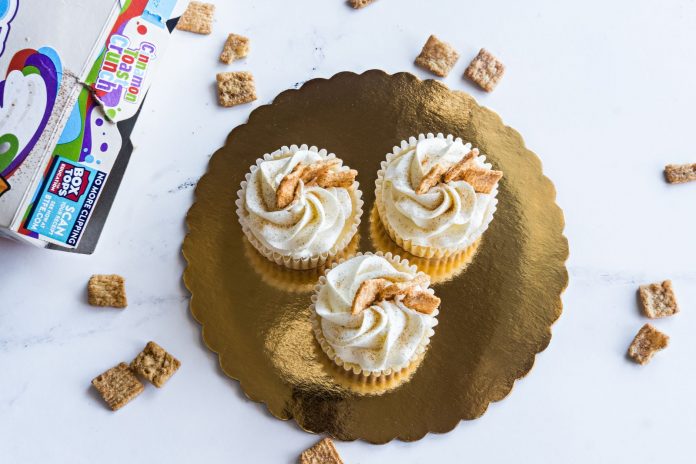 Top view of three cinnamon toast cupcakes with frosting on a golden tray, surrounded by cereal.