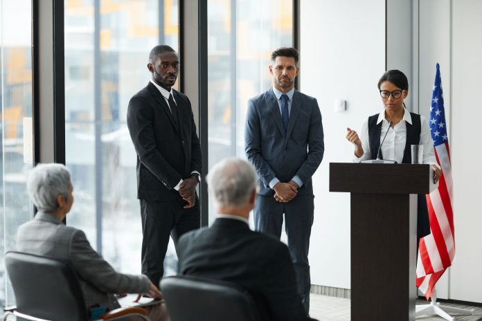mayor-solomon-urges-school-board-to-reject-superin Professional speakers presenting at a conference in a modern room with glass windows.