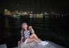 Woman sitting by the waterfront at night with city lights in the background.