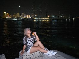 Woman sitting by the waterfront at night with city lights in the background.
