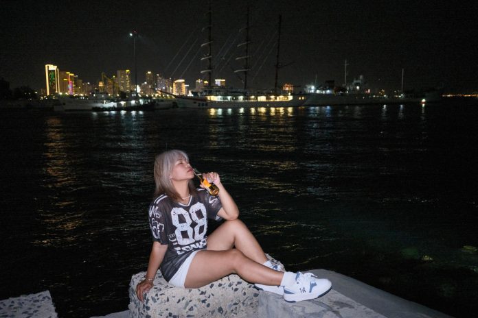 Woman sitting by the waterfront at night with city lights in the background.