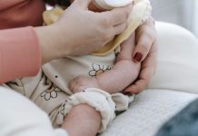 Crop anonymous mother embracing and feeding adorable newborn baby with milk from bottle at home