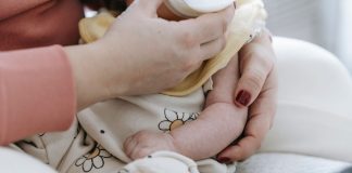 Crop anonymous mother embracing and feeding adorable newborn baby with milk from bottle at home