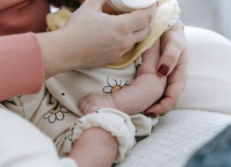 Crop anonymous mother embracing and feeding adorable newborn baby with milk from bottle at home