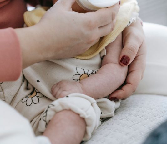Crop anonymous mother embracing and feeding adorable newborn baby with milk from bottle at home