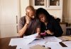 A couple sits at a table managing domestic finances, evaluating documents and using a smartphone.