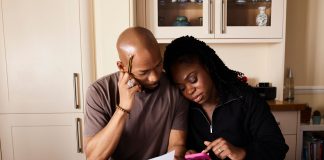 A couple sits at a table managing domestic finances, evaluating documents and using a smartphone.