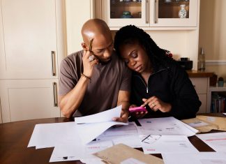 A couple sits at a table managing domestic finances, evaluating documents and using a smartphone.