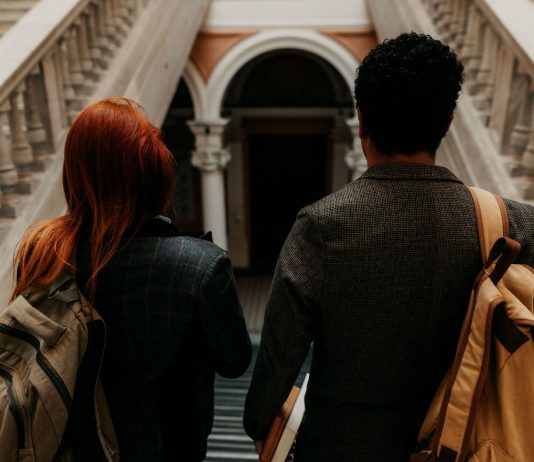 Two university students with backpacks walk down a grand staircase in a historic building.