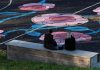 This July 4th, Art in Liberty State Park Will Be Invisible — Until You Look Through Your Phone Two women sit on a bench facing a sports field with colorful floral designs in Jönköping, Sweden.