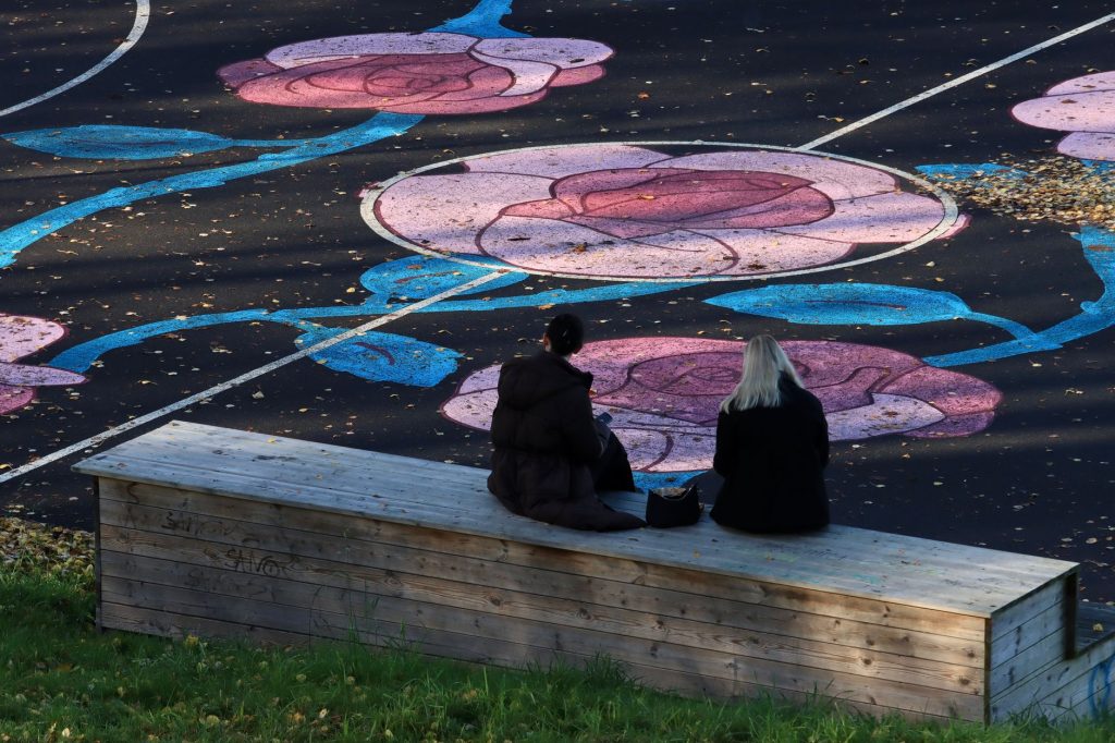 Two women sit on a bench facing a sports field with colorful floral designs in Jönköping, Sweden.