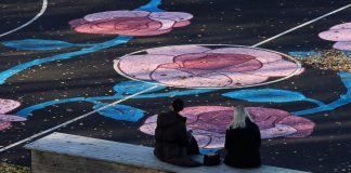This July 4th, Art in Liberty State Park Will Be Invisible — Until You Look Through Your Phone Two women sit on a bench facing a sports field with colorful floral designs in Jönköping, Sweden.