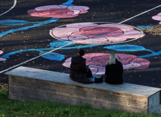 Two women sit on a bench facing a sports field with colorful floral designs in Jönköping, Sweden.