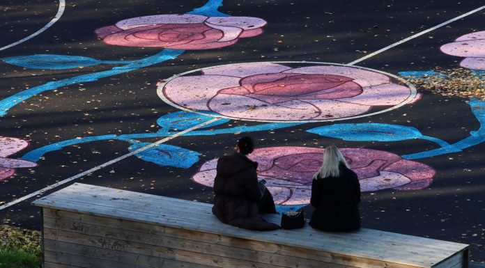 Two women sit on a bench facing a sports field with colorful floral designs in Jönköping, Sweden.
