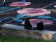 This July 4th, Art in Liberty State Park Will Be Invisible — Until You Look Through Your Phone Two women sit on a bench facing a sports field with colorful floral designs in Jönköping, Sweden.