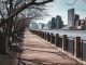 Bare trees line a waterfront walkway in New York City, with skyscrapers in the background.