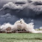 Powerful waves crash against a pier and lighthouse under dark stormy skies.