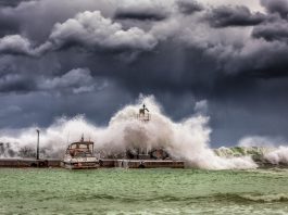 Powerful waves crash against a pier and lighthouse under dark stormy skies.