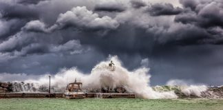 Powerful waves crash against a pier and lighthouse under dark stormy skies.