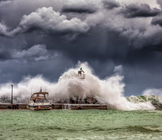 Powerful waves crash against a pier and lighthouse under dark stormy skies.