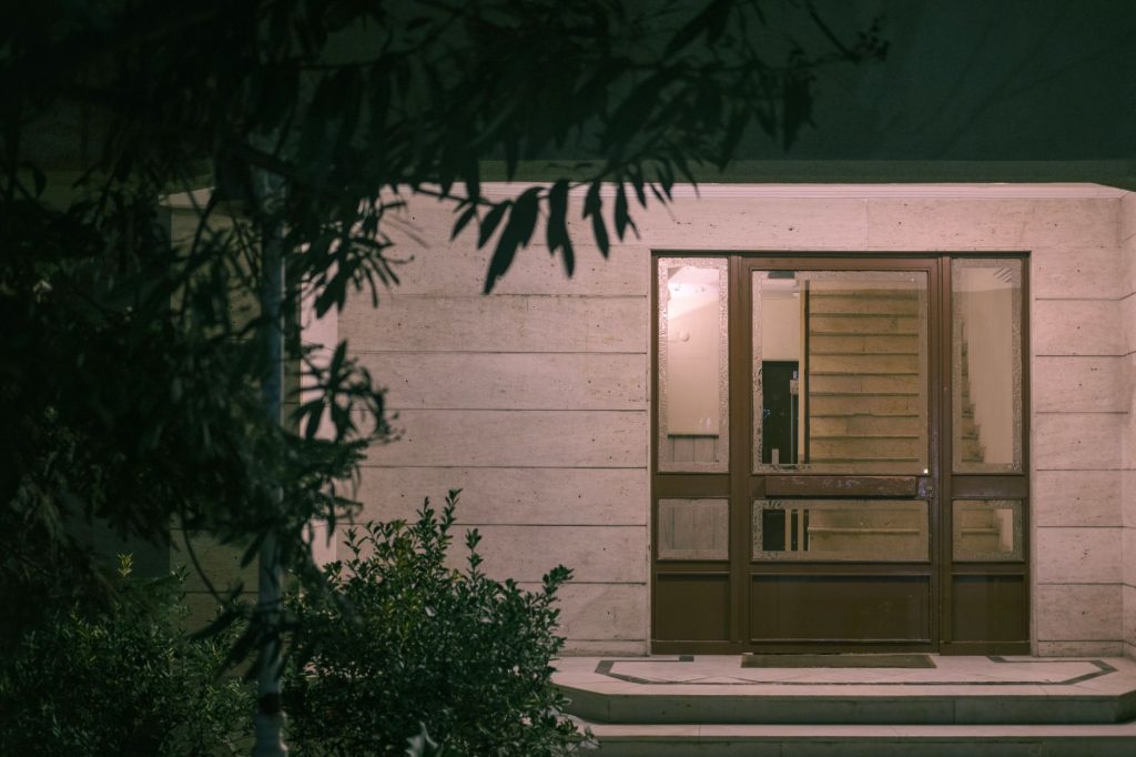 A serene nighttime view of a modern building entrance with surrounding foliage, creating a warm and inviting atmosphere.