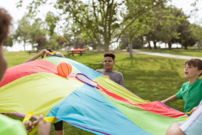 autism-summer-camp-gives-hudson-families-a-respite Kids enjoying a fun outdoor game with a colorful parachute and ball in a park setting.
