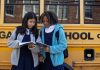 Multiethnic schoolchildren in uniforms watching exercise book near yellow school bus reflecting building in town