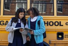 Multiethnic schoolchildren in uniforms watching exercise book near yellow school bus reflecting building in town
