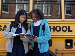 Multiethnic schoolchildren in uniforms watching exercise book near yellow school bus reflecting building in town