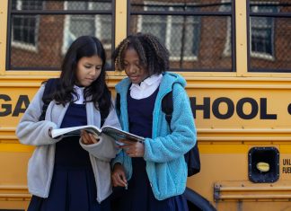 Multiethnic schoolchildren in uniforms watching exercise book near yellow school bus reflecting building in town