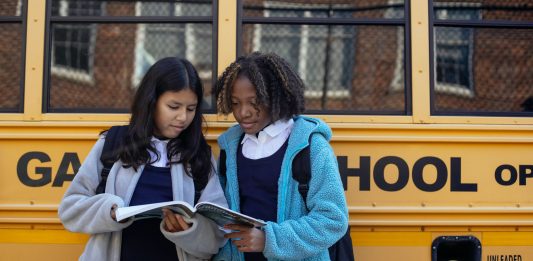 Multiethnic schoolchildren in uniforms watching exercise book near yellow school bus reflecting building in town