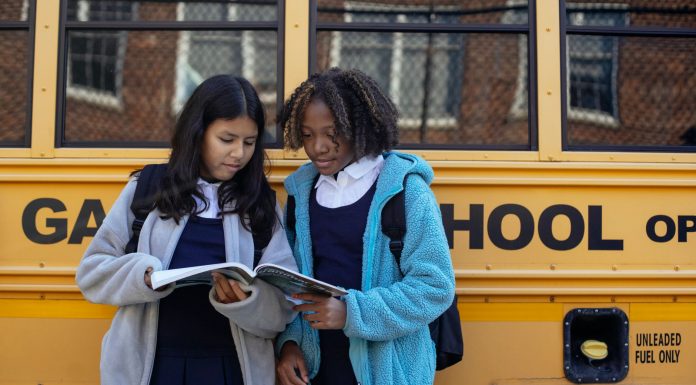 Multiethnic schoolchildren in uniforms watching exercise book near yellow school bus reflecting building in town
