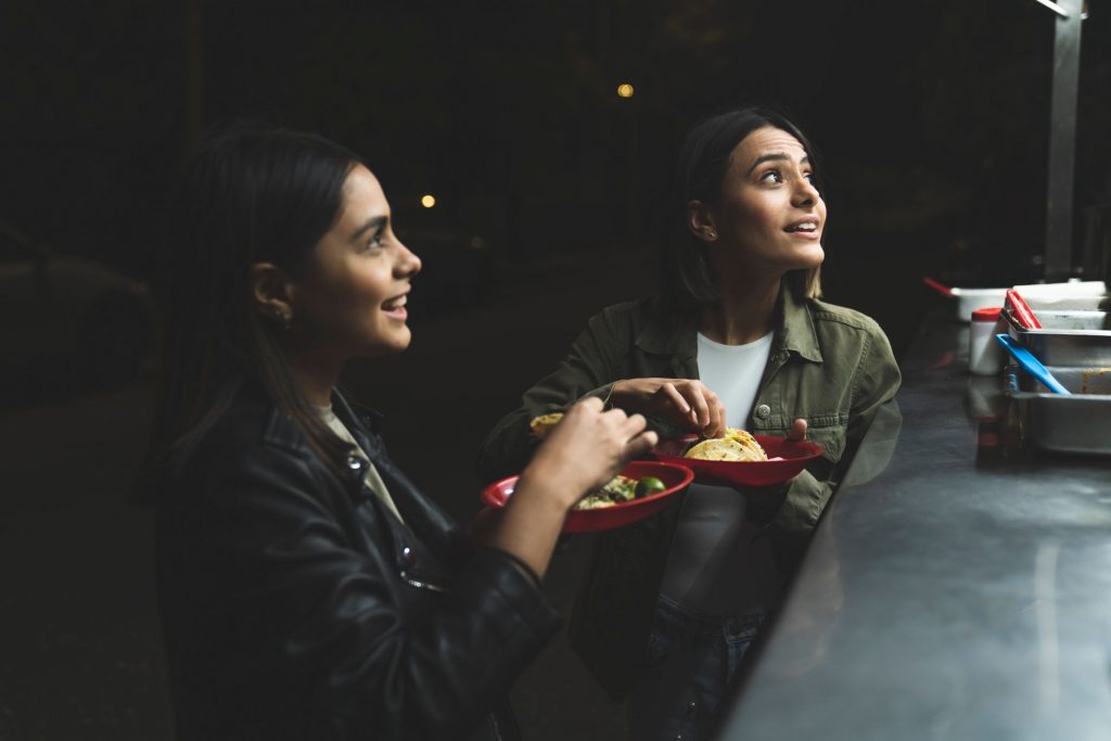 Two women joyfully ordering tacos at a vibrant nighttime street stand.
