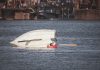 A capsized boat near an urban waterfront with a person wearing a life jacket visible in calm waters.