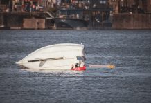 A capsized boat near an urban waterfront with a person wearing a life jacket visible in calm waters.