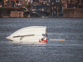 A capsized boat near an urban waterfront with a person wearing a life jacket visible in calm waters.